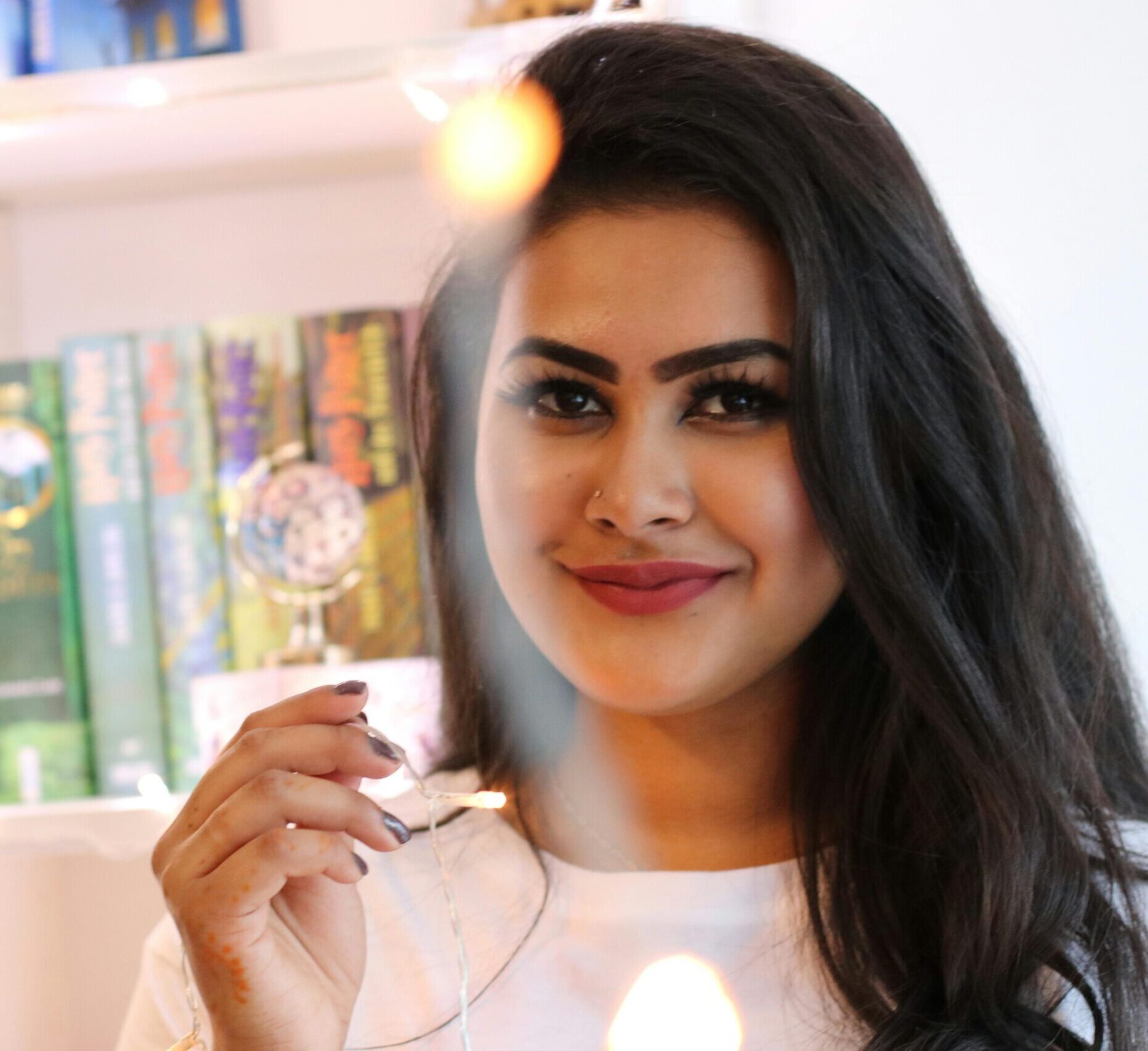 Portrait of a smiling woman holding string lights indoors by bookshelf.